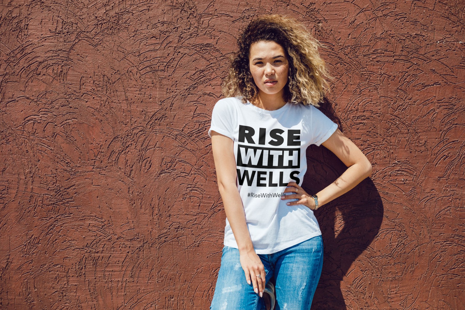 A woman wearing the Rise With Wells tee against a rust-red wall